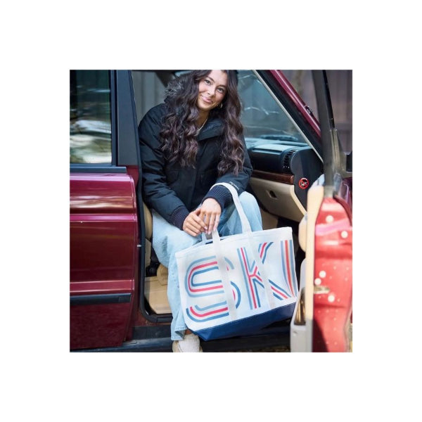 A woman sitting in the open door of a red car holds a large white tote bag with bold red and blue striped lettering that reads 'SK'.