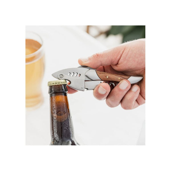 A close-up of a hand using a metal and wood bottle opener to remove a gold cap from a brown glass beer bottle, with a blurred glass of beer in the background.