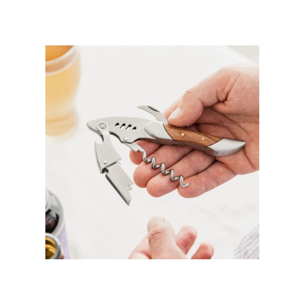 A close-up of a hand holding a stainless steel corkscrew with a wooden handle, shown against a softly blurred kitchen background with a glass of beer and a bottle partially visible.