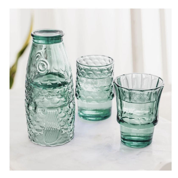 A set of three translucent light green glassware pieces, including a textured bottle and two matching tumblers, arranged on a white marble surface against a soft white curtain background.