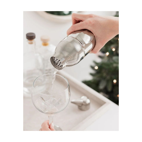 A stainless steel cocktail shaker being used to pour a clear liquid into a wine glass, with a hand holding the shaker and another hand holding the glass. The background is softly blurred with a holiday tree and warm lights.