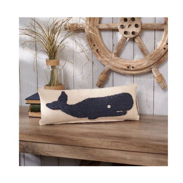 A rectangular beige throw pillow with a dark navy blue whale silhouette, styled on a wooden table with a rustic nautical ship's wheel and dried grass in the background.